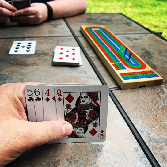 Cribbage table with Cribbage board. Board shows each player a little over 2/3 of the way to game.
Foregrounded is one player's hand (body part) and hand (cards: 5♣, 6♣, 4♦, Q♦)
Midground is 7♠ in play and 8♦ as starter.
Background is other player's left arm and both hands (body parts) holding hand (cards)