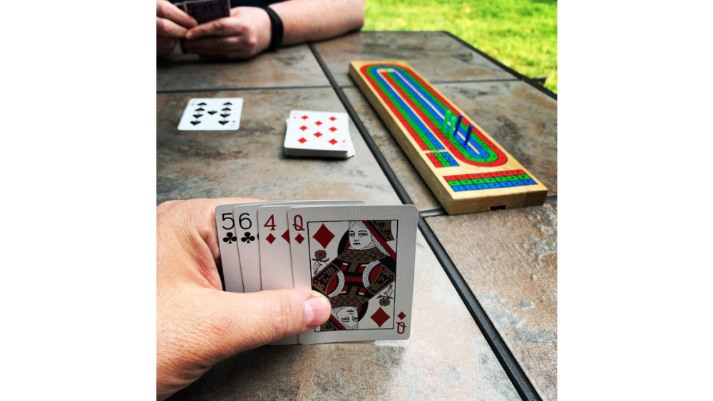 Cribbage table with Cribbage board. Board shows each player a little over 2/3 of the way to game. Foregrounded is one player's hand (body part) and hand (cards: 5♣, 6♣, 4♦, Q♦) Midground is 7♠ in play and 8♦ as starter. Background is other player's left arm and both hands (body parts) holding hand (cards)